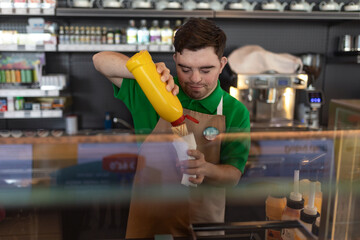 Happy waiter with Down syndrome standing by counter and prepairing hot-dog to a customer in cafe at...