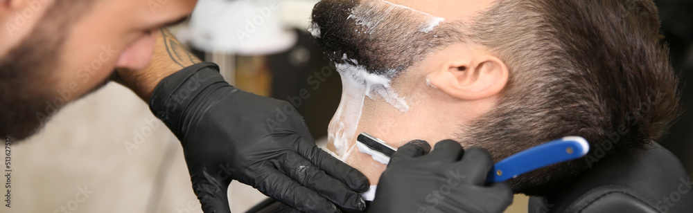 Wall mural Professional hairdresser shaving client with straight razor in barbershop, closeup. Banner design