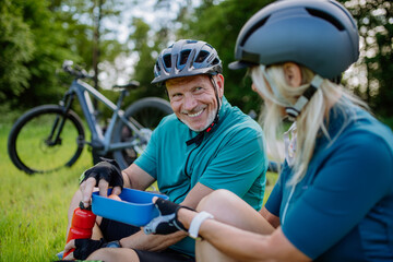 Obraz premium Active senior couple resting after bicycle ride at summer park, sitting on grass and having snack.