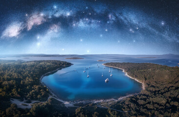 Milky Way arch and beautiful yachts and boats on the sea bay at summer night. Kamenjak, Croatia. Space. Top view of arched milky way, starry sky, sailboats, lagoon, clear blue water, forest. Travel