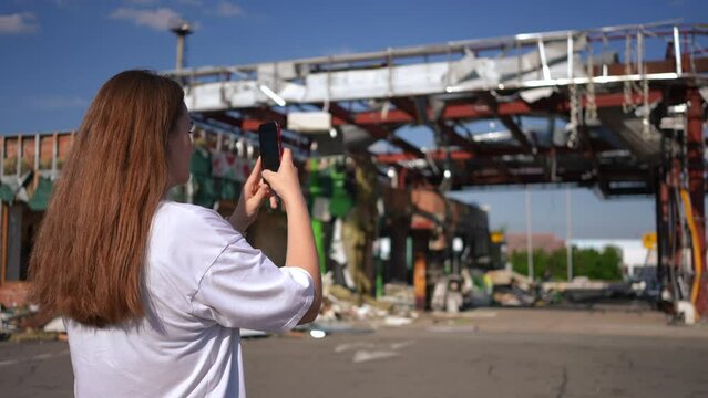 Back view young woman taking photos of ruined gas station in Kyiv Ukraine. Shooting over shoulder of brunette Ukrainian lady photographing consequences of bombing in slow motion