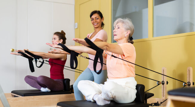 Positive senior woman doing stretching exercises on pilates reformer as part of injury rehabilitation course. Therapeutic physical training concept