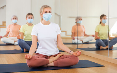 Portrait of blonde woman in face mask making yoga meditation in lotus pose in fitness studio
