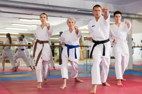 Various aged men and women in white kimono and belts training kata moves in gym.