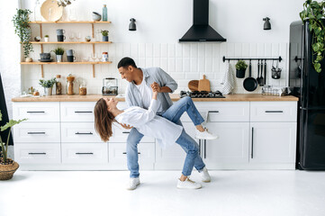 A young modern happy couple in love, newlyweds, stylishly dressed, dance together to their favorite music at home in the kitchen with stylish interior, smile, enjoying to spending time with each other