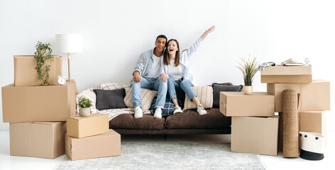 Panoramic photo of excited happy multiracial young couple in love, sitting on sofa in living room of their new home between cardboard boxes, they invested in real estate, happy with purchase, smiling