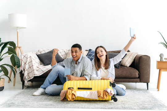 Cheerful multiracial couple, packed a trip suitcase for vacation, sit on the floor in living room near the yellow suitcase, hold their passports, look at camera, smile happily. Long-awaited vacation - Powered by Adobe