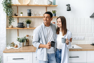 Happy multiracial romantic couple, newlyweds, stylishly dressed, stand hugging at home in the kitchen, hold cups of coffee or tea in their hands, look away, dream, smile, spend morning time together