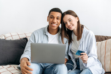 Online shopping, technology. Positive multiracial couple in love, spouses, sitting on sofa in a living room, using laptop and credit card for online shopping, delivery, looking at camera, smiling