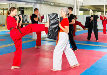 European young woman exercising kicks with senior woman who holding punching pad during group karate training.