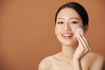 Joyful young Vietnamese woman wiping face with cotton pad soaked in toner