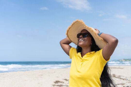 Cheerful Young Female In Summer Hat And Sunglasses Touching Head And Looking Away With Smile While Relaxing Against Sea On Sunny Weekend Day