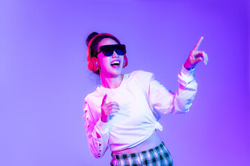 Young asian woman in white fashion crop sweatshirt wearing sunglasses and red headphones listen to music dancing and finger pointing on the purple background.
