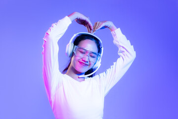 Cheerful Young asian woman in white shirt wearing headphones posing love hands symbol on blue and purple background.
