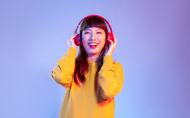 Young asian woman in yellow sweatshirt wearing red headphones smiling and listen to music on the purple screen background.
