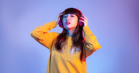 Young asian woman in yellow sweatshirt wearing red headphones listen to music on the purple screen background.
