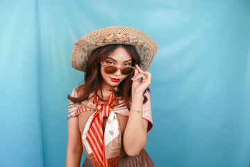 Young Asian confident and happy woman is glancing and holding her sunglasses isolated by a blue background.