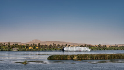 There are islands with green vegetation in the riverbed. A white cruise ship is sailing on blue water. A house is visible among the palm trees on the shore. Sand dunes against the sky. Egypt. Nile