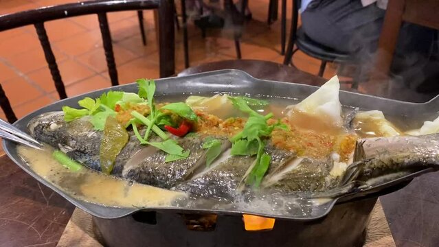 Thai style garlic lime steam sea bass barramundi fish on hot fish serving platter burner flame hot steam bubbling
