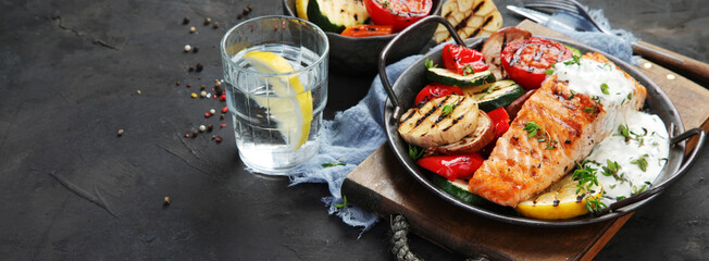 Salmon steak with vegetables and white sauce on dark background.
