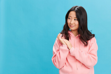 Happy lovely Asian student young lady in pink hoodie sweatshirt smiling posing isolated on over blue studio background. The best offer for ad. People Emotions for Everyday concept