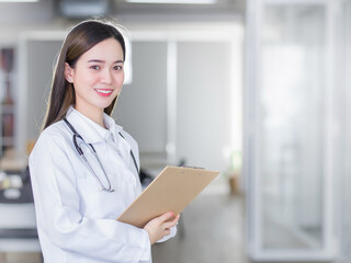Professional beautiful young female doctor holding document in clipboard smiling looking at the camera while she wears a white lab coat and a stethoscope in hospital.