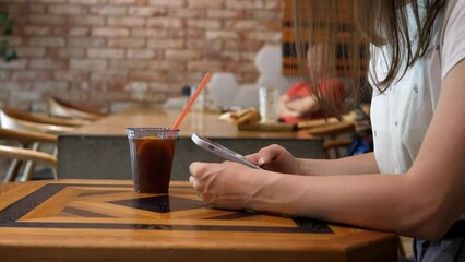 Woman, in a cafe, is waiting for an order, holding a mobile phone in her hand, typing, scrolling through the page using the application. Internet news app content concept of mobile phone dependency.