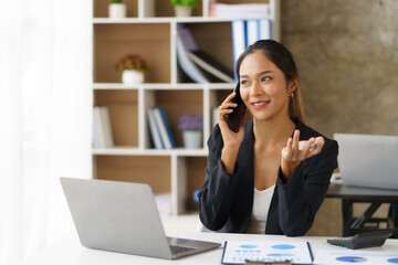 Happy young Asian business woman sitting smiling and talking on mobile phone in office.