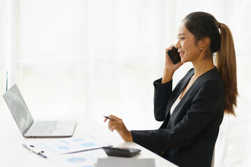 Happy young Asian business woman sitting smiling and talking on mobile phone in office.