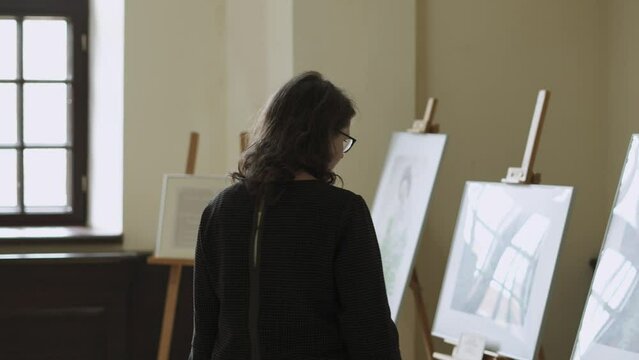 Young thoughtful woman in glasses and plaid dress goes to paintings at the exhibition and looks at picture. Lady visits art gallery in medieval castle, museum.