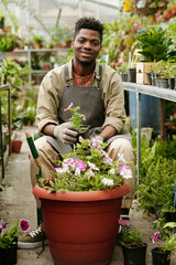 Portrait of African young gardener smiling at camera while sitting in flower shop and making...