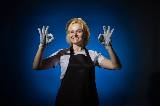 A woman in a white t-shirt, black apron, gloves and a hat smiles and shows an OK sign with her fingers on a dark background.