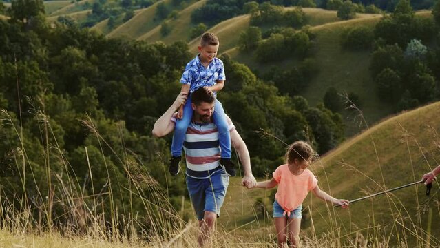 Happy father carries his son on his shoulders having a nature trip with his family in hills