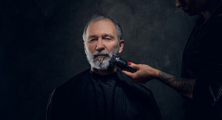 Shot of female barber with tattooed hand cutting elderly man using clipper against dark background.