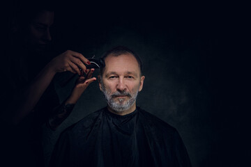 Portrait of cool elderly man being cutting by female barber against dark background.