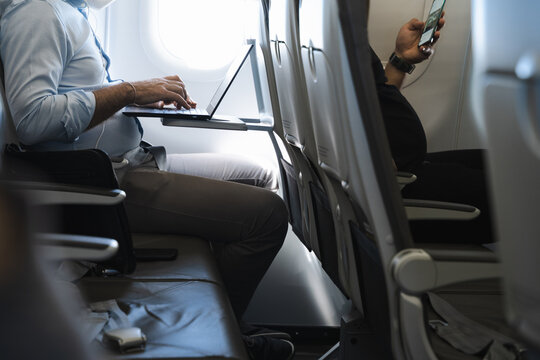 Businessman using laptop in airplane and passenger in front row with cellphone. Airline wifi connection, device flight mode concepts