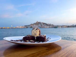 Selective focus on foreground on Chocolate brownie with ice cream in a romantic restaurant in the new Marina Botafoch in Ibiza. Sea and Ibiza old town in the blurred background. 