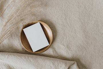 Flatlay of blank paper card on brass dish, pampas grass on neutral beige crumpled linen cloth....
