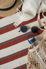 Fashion composition with women's accessories on colorful blanket. String bag, straw hat, sunglasses, bottle of water. Minimal lifestyle concept