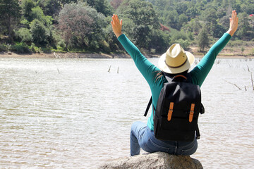 Latin adult woman with hat and sunglasses sitting on a stone next to a lake makes the victory sign...