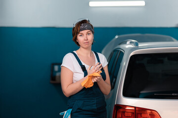 Portrait of a caucasian young woman in a blue coveralls waping her hands with a rag. Car and...