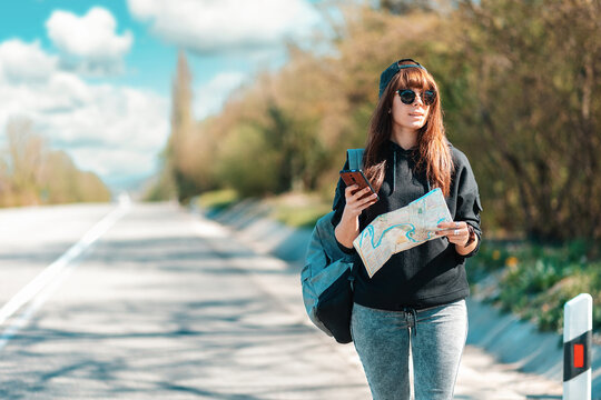 Local Travel and hitchhiking. Pretty young woman in a cap and sunglasses, holding a paper map. Road is blurred. Copy space