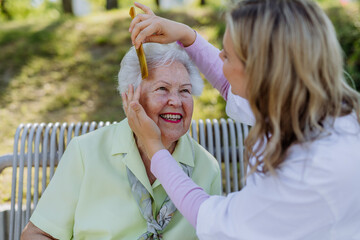 Caregiver helping senior woman to comb hair and make hairstyle when sitting on bench in park in summer. - obrazy, fototapety, plakaty