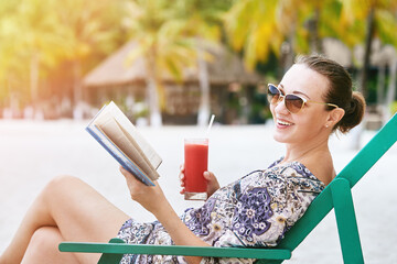 Happy smiling woman with book and glass of juice relaxing in sun lounger on beach in hotel.