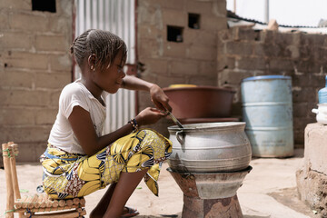 Small African girl sitting in front of a simple charcoal stove intent on mixing the contents of a...