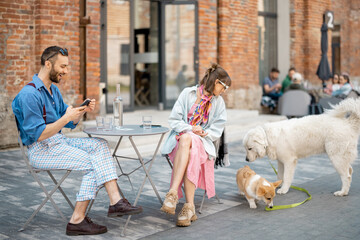 Stylish couple sit at cafe while dogs playing around in modern office district. Leisure time with...