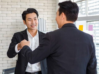 Happy cheerful Asian millennial professional successful male entrepreneur and unrecognizable businessman in formal suit standing together holding hands fists bumping celebrating job agreement done.