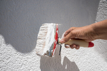 womans hand with paint brush, saturated with white dispersion paint. renovating the wall outdoor