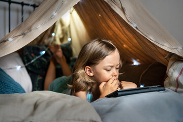 Happy cute little 6 year old boy having fun playing in teepee tent. Child using digital tablet watching cartoons or playing computer games lying in kid tent at home. 