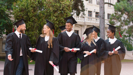 Modern college park group of multiracial graduates students after the graduation walking in front of the camera and discussing holding diplomas smiling large and feeling excited